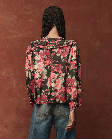 A person with braided hair wearing The Symphony Top in Charming Rose from THE GREAT. HOL 24, paired with jeans, stands against a textured brown background.