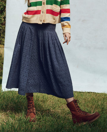 A person in a striped sweater and brown lace-up boots stands gracefully on the grass, wearing THE GREAT. PS25's The Rhythm Skirt in Riverside Blue that sways with every step.
