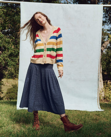 A person stands on grass, embodying fashion in a striped cardigan, The Rhythm Skirt. -- Riverside Blue from THE GREAT. PS25, and brown boots, set against a light blue backdrop with trees in the background.