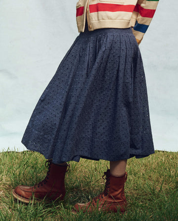 On the grass, a person elegantly displays their style in a striped sweater, chic Riverside Blue Rhythm Skirt from THE GREAT. PS25, and brown lace-up boots.