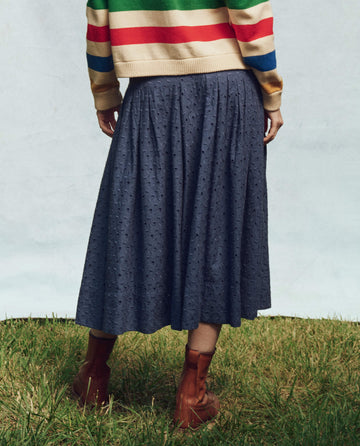A fashion-forward individual wearing a striped sweater, The Rhythm Skirt in Riverside Blue by THE GREAT. PS25, and brown ankle boots stands gracefully on the grass.