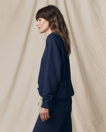 A woman with long brown hair stands in profile against a beige fabric background, wearing THE GREAT. FALL 23 KNITS The College Sweatshirt in Solid -- True Navy with matching pants.