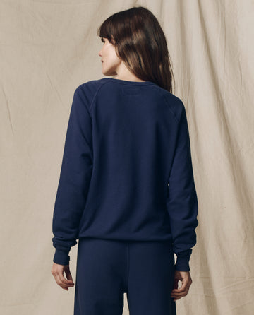 A person with long brown hair wears THE GREAT. FALL 23 KNITS The College Sweatshirt, Solid in True Navy, with matching pants, standing with their back to the camera against a beige fabric backdrop.