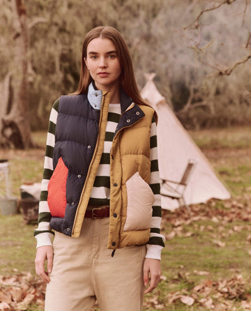 A person wearing The Down Polar Vest in Appalachian Colorblock from THE GREAT. FALL 24 TGO stands outdoors near a tree and a white tent. Fallen leaves cover the ground, creating an ideal setting for adventure accessories.