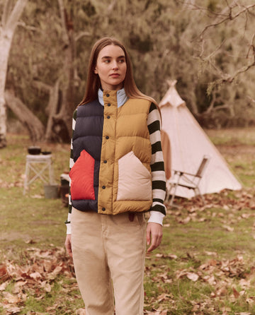 A woman is outdoors in a park, surrounded by trees and a tent in the background, wearing THE GREAT. FALL 24 TGO's The Down Polar Vest in Appalachian Colorblock along with a striped shirt.