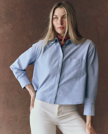 A woman stands against a brown background wearing THE GREAT. F25 D1's The Cropped Oxford Shirt in Classic Blue, styled with white pants and a red bandana around her neck.