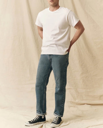 A person wearing THE GREAT. FALL 23 MEN's The Men's Pure Knits Boxy Crew in True White, blue jeans, and black high-top sneakers stands against a beige fabric backdrop.