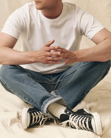 Wearing THE GREAT. FALL 23 MEN's The Men's Pure Knits Boxy Crew in True White, blue jeans, and black sneakers, a person sits cross-legged on a light surface with hands clasped.