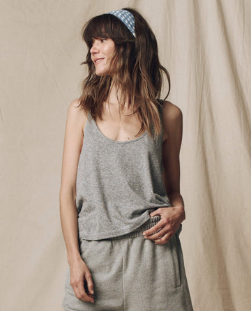 A woman with loose hair and a blue polka-dot headband smiles to the side, wearing THE GREAT. SU25 CORE KNITS' The Scoop Neck Tank in Heather Grey, styled with shorts against a beige fabric backdrop.