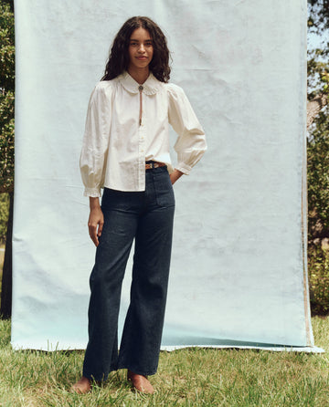 A person stands outdoors on grass in front of a backdrop, wearing a stylish white Hemingway Top by THE GREAT. PS25 and dark jeans.
