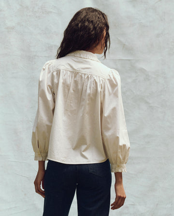 A person with long hair, wearing The Hemingway Top in white by THE GREAT. PS25, and dark pants, stands with their back to the camera against a neutral background.