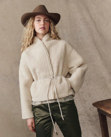 A person wearing THE GREAT. H25 COLLECTION’s The Reversible Drover Coat in Malt, a brown wide-brimmed hat, and dark green satin pants stands in front of a beige backdrop.