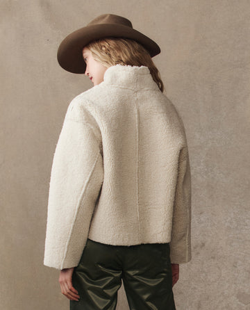 A person in a brown hat, dark green pants, and THE GREAT. H25 COLLECTION's The Reversible Drover Coat in Malt faces away from the camera against a neutral background.
