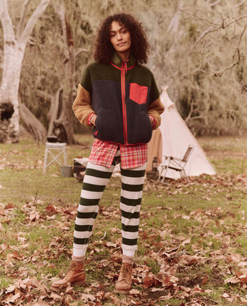 A person revels in an everyday adventure, wearing The Union Long John in Mountain View Stripe from THE GREAT. FALL 24 TGO collection on leafy ground. The scene includes a tent and trees, perfect for showcasing their technical clothing designed for both style and the great outdoors.