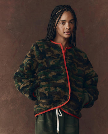 A person with braided hair wears THE GREAT. F25 D1 "The Reversible Plush Bomber – Camo & Hot Red" with green pants, standing against a brown background.
