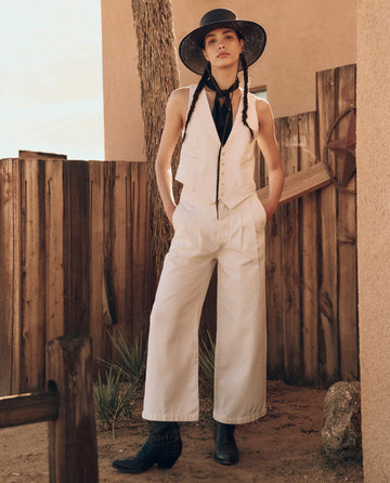 A person wearing THE GREAT. PF24's The Suit Vest -- Natural Hemp paired with white pants, a black hat, and black boots stands next to a wooden fence in an outdoor setting.