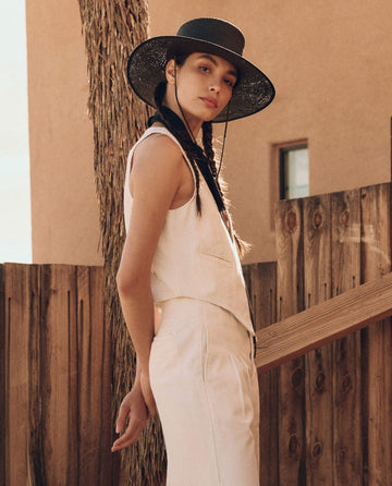 Woman in The Suit Vest by THE GREAT. PF24, a natural hemp sleeveless top and pants ensemble, wearing a black wide-brimmed hat, standing near a wooden fence and building with her head slightly tilted and a neutral expression. The casual elegance of her cotton hemp denim clothing complements the rustic backdrop perfectly.