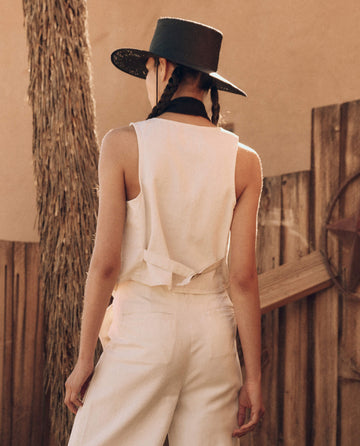 A person with braided hair wearing a black hat and a sleeveless white outfit reminiscent of THE GREAT. PF24's The Suit Vest - Natural Hemp, stands outdoors facing away from the camera against a rustic wooden background.