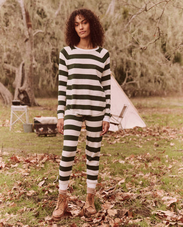 Dressed in The Union Long John by THE GREAT. FALL 24 TGO, featuring the Mountain View Stripe design, a person stands on grass sprinkled with fallen leaves, near a tent and camping gear amidst trees, poised for an everyday adventure.