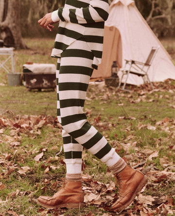 Dressed in The Union Long John – Mountain View Stripe by THE GREAT. FALL 24 TGO, paired with brown boots, a person strolls on the grass with a tent and chair in the background, perfectly embodying the spirit of an everyday adventure.