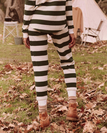 Dressed in The Union Long John in Mountain View Stripe by THE GREAT. FALL 24 TGO, a person stands on fallen leaves near a tent and camping gear, ready for their everyday adventure in brown boots.