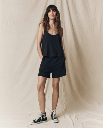 A woman stands against a beige fabric backdrop wearing a black tank top, THE GREAT. SU25 CORE KNITS The Victory Short. Solid -- Almost Black, and black high-top sneakers.