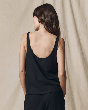 A person with long brown hair stands facing away, wearing THE GREAT. SU25 CORE KNITS The Scoop Neck Tank in Solid -- Almost Black, paired with black pants, in front of a neutral fabric background.