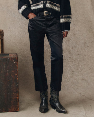 Wearing THE GREAT. H25 COLLECTION’s The Satin Wayne pants in black, paired with a black patterned sweater, a statement belt, and black cowboy boots, a person stands by a wooden box against a neutral backdrop.