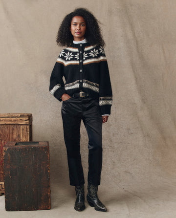 A woman poses before a beige backdrop, wearing THE GREAT. H25 COLLECTION's The Satin Wayne in black, paired with black pants, a belt, and black boots. Wooden boxes sit beside her, while satin accents elevate her look.