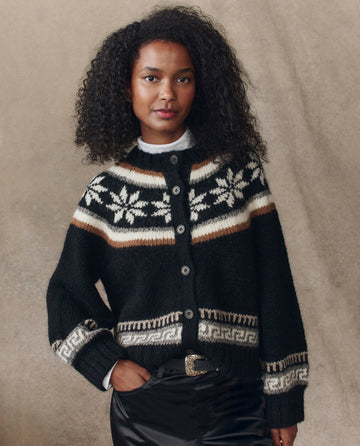 A woman with curly hair models THE GREAT. H25 COLLECTION’s The Snowflake Ice Rink Cardigan in black over a white collared shirt, standing against a neutral backdrop.