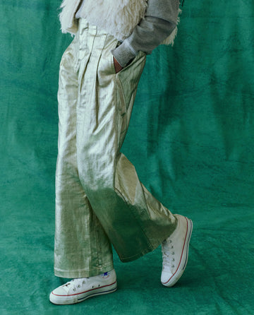 A person stands against a green background, wearing The Sculpted Trouser in Starlight from THE GREAT. HOL 24, featuring metallic foil-coated wide-leg pants with a high-waisted silhouette. They pair it with white high-top sneakers and a gray top accented by a chic fur vest.