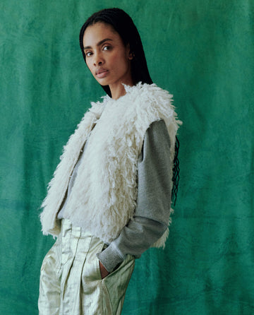 A person wearing The Boucle Loop Vest in Soft White by THE GREAT. HOL 24 over a gray sweater and shiny pants stands against a vibrant green background.
