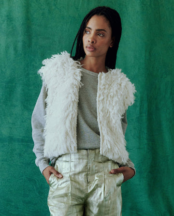 A person wearing THE GREAT. HOL 24's soft white 'The Boucle Loop Vest', a gray sweater, and metallic pants stands against a green background.