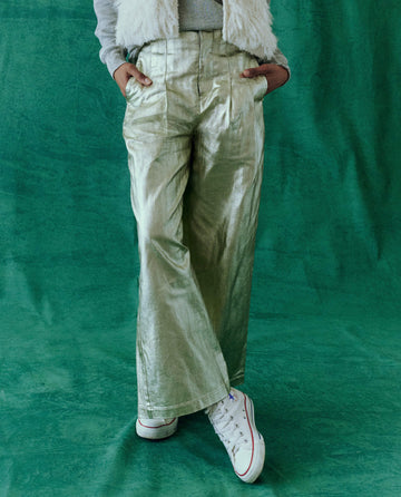 Against a green background, the person is wearing THE GREAT. HOL 24's Starlight Sculpted Trouser, crafted from metallic foil-coated twill, paired with white sneakers.