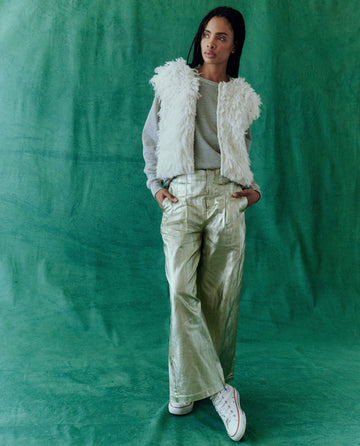 A person in the Sculpted Trouser from THE GREAT. HOL 24, featuring a metallic foil-coated finish and a high-waisted silhouette, pairs it with a textured vest and gray sweater while standing against a green backdrop, hands in pockets.