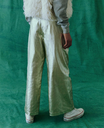 A person stands against a green backdrop, flaunting "The Sculpted Trouser - Starlight" by THE GREAT. HOL 24 in a high-waisted silhouette. Complemented by a gray shirt and white fluffy vest, the ensemble is completed with white sneakers.