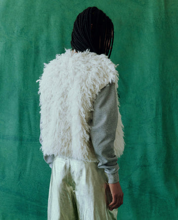 A person with braided hair stands against a green background, wearing The Boucle Loop Vest in Soft White from THE GREAT. HOL 24, paired with a gray long-sleeve shirt and shiny light-colored pants.