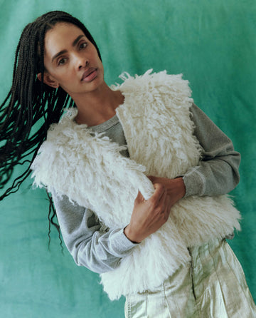 Wearing The Boucle Loop Vest in soft white from THE GREAT. HOL 24 over a gray sweater, a person with long braids stands against a lush green background.