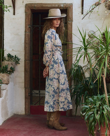 A woman in THE GREAT. PF25 The Pampas Dress – Indigo Floral, with a wide-brimmed hat and brown boots, stands sideways on a red-tiled floor near potted plants in front of a rustic doorway.