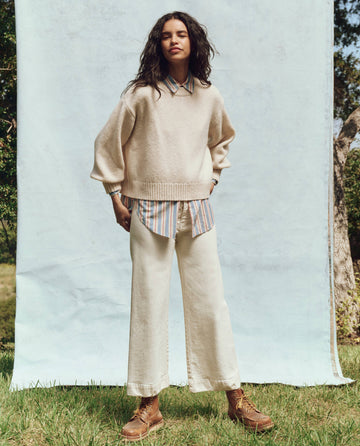 In front of a light blue backdrop, a person stands outdoors wearing a cream sweater over a striped shirt paired with THE GREAT. PS25's The Seafair Jean in Vintage Stone, featuring ultrahigh waist and wide legs, alongside brown boots.