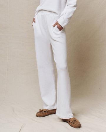 A person in a white lounge set, with hands in pockets and wearing brown moccasins, stands on a light fabric backdrop. The Victory Sweatpant in True White from THE GREAT. SP25 POLO LOUNGE collection embodies the elegance of premium activewear.