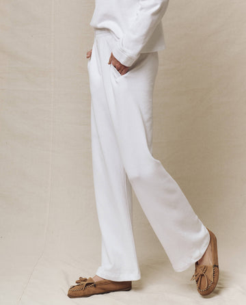An individual in a white long-sleeve top, The Victory Sweatpant in True White by THE GREAT. SP25 POLO LOUNGE, and brown moccasin-style shoes stands against a plain backdrop.