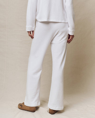 A person wearing a white long-sleeve top and THE GREAT. SP25 POLO LOUNGE Victory Sweatpant in True White stands on a neutral fabric backdrop with tan moccasin-style shoes.