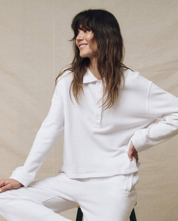 A person with long hair, wearing THE GREAT. SP25 POLO LOUNGE's The Rugby Sweatshirt in True White and pants, sits on a stool against a beige background.