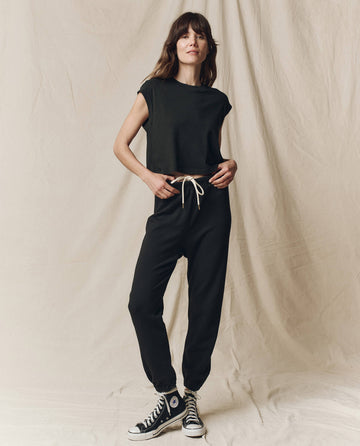 A person stands on a fabric-draped floor, wearing a black sleeveless top and The Stadium Sweatpant in Almost Black by THE GREAT. CORE KNITS, featuring an elasticated waist with a white drawstring, paired with black high-top sneakers.