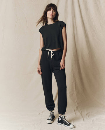 Person in front of a beige fabric backdrop wearing THE GREAT. SU23 CORE KNITS' The Square Tee in Almost Black, with matching jogger pants and black high-top sneakers.