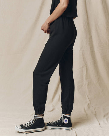 A person stands on a beige fabric background wearing THE GREAT. CORE KNITS' The Stadium Sweatpant in Solid Almost Black and black high-top sneakers, highlighting its elasticated waist for comfort.