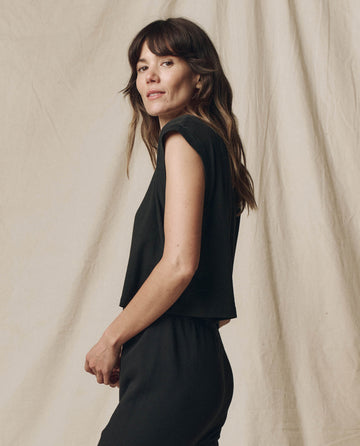 A person with long hair stands sideways, wearing The Square Tee in Almost Black by THE GREAT. SU23 CORE KNITS against a beige fabric backdrop, reminiscent of a vintage athletic style.