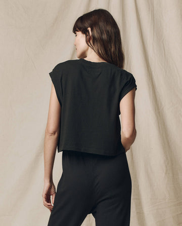 A person with long hair in The Square Tee by THE GREAT. SU23 CORE KNITS in Almost Black, standing against a neutral fabric backdrop, exuding the vibe of a vintage athletic tee.