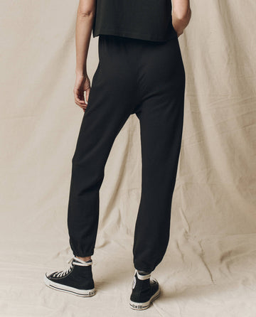 A person in chic black joggers, resembling THE GREAT. CORE KNITS' The Stadium Sweatpant in Almost Black, paired with sneakers, stands on a beige fabric background. These joggers have an elastic waistband for comfort and are made from 100% cotton, providing an ideal mix of style and ease.